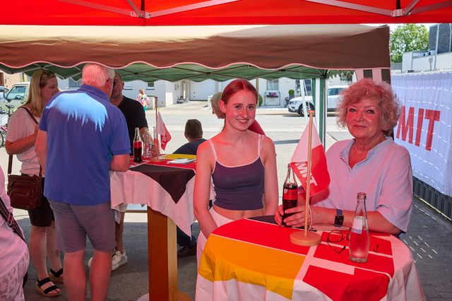Zwei Frauen lachen unter rot-weißem Sonnenschirm auf Markt mit bunten Tischdecken.