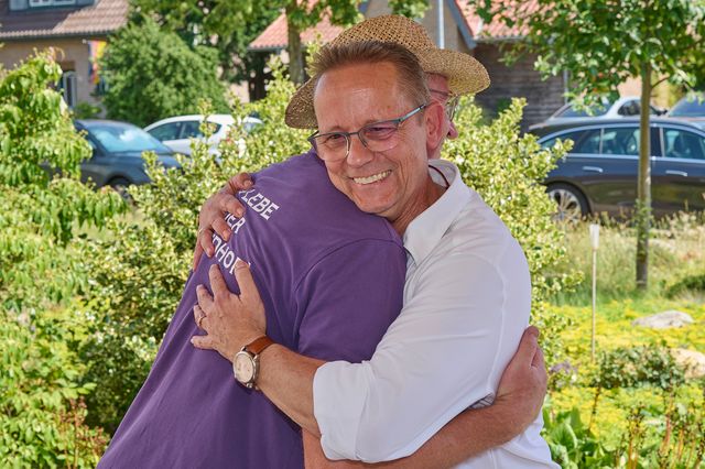 Glücklicher Mann mit Blumenkrone umarmt Person in lila Shirt herzlich im Garten.