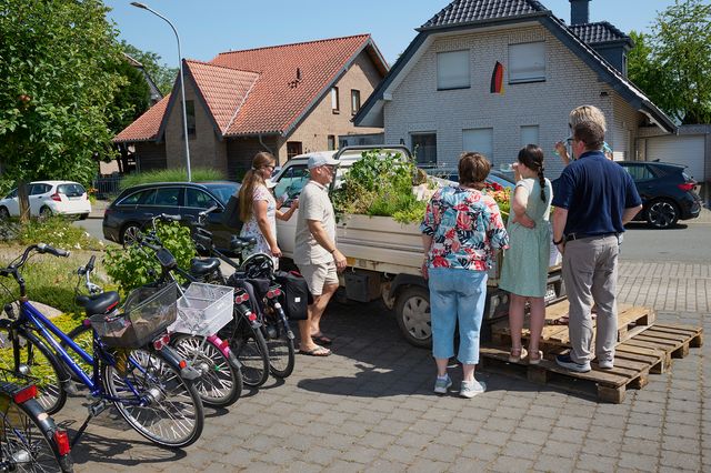 Gruppe von Menschen mit Fahrrädern vor einem Haus mit Blumenbeeten und Traktor.