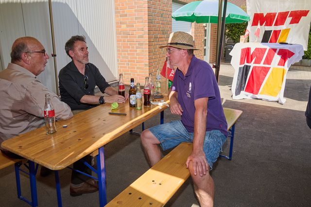 Drei Männer sitzen draußen an Tisch mit Bier und unterhalten sich bei sonnigem Wetter.