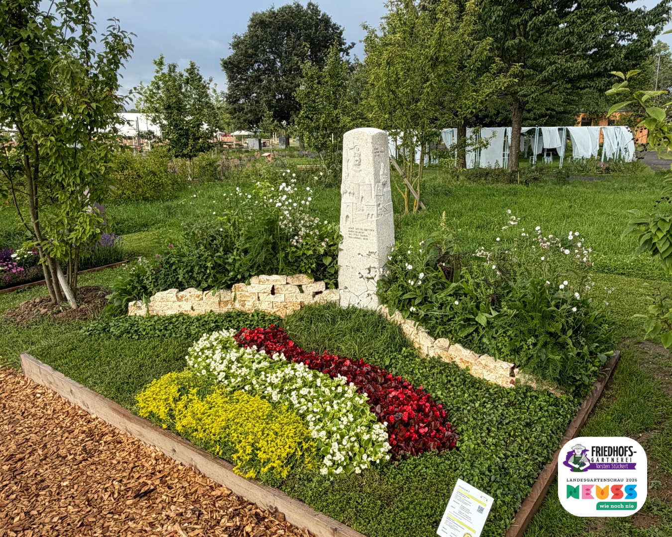 Blumengestalteter Gedenkstein in gepflegter Grünanlage mit farbenfrohen Blumenbeeten.
