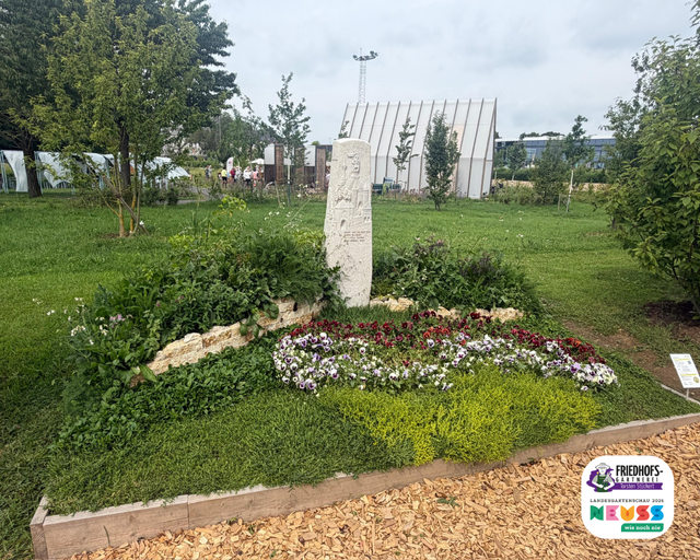 Gepflegter Gedenkstein mit Blumenbeet und grünem Rasen in parkähnlicher Umgebung.