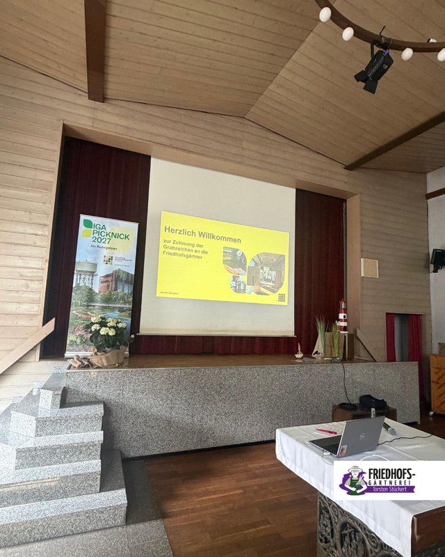 Vortragsraum mit Präsentation zu „Herzlich Willkommen“ auf Bühne, Projektor und Podium.