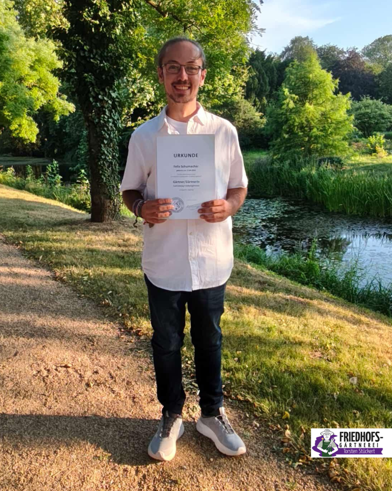 Glücklicher Mann hält Urkunde in parkähnlicher Umgebung mit Wasserlauf.